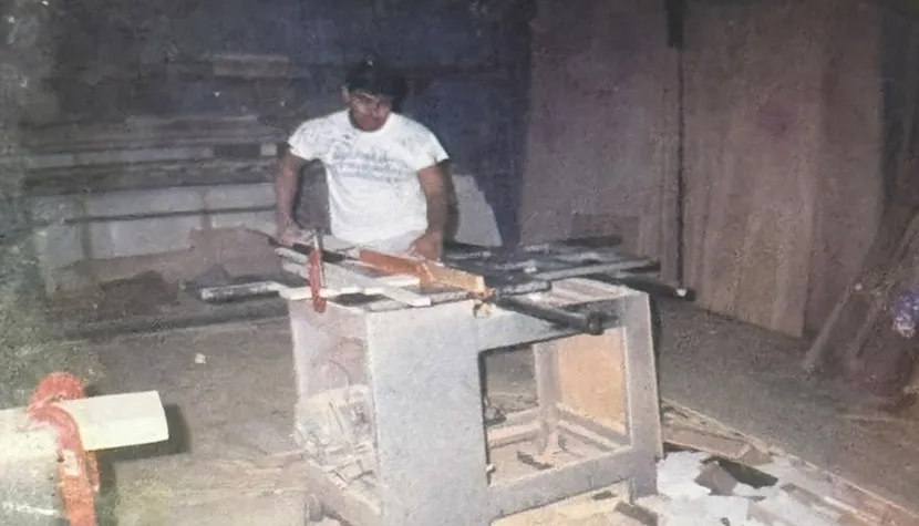 Fotografia antiga de um marceneiro trabalhando em uma máquina, com pranchas de madeira ao fundo