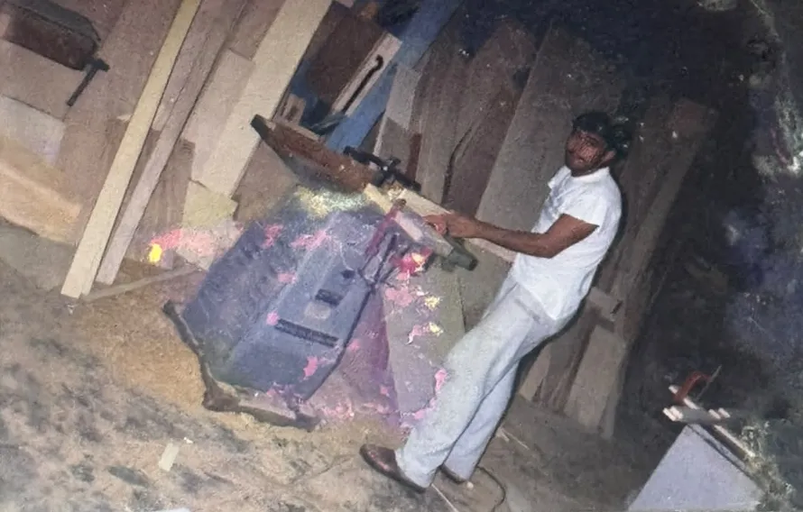 Fotografia antiga de um marceneiro operando uma máquina, com serragem no chão e tábuas encostadas