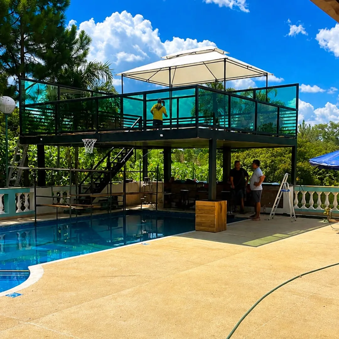 Deck elevado com guarda-corpo de vidro verde ao lado de uma piscina, sob céu azul