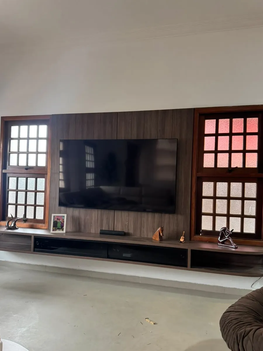 Painel de TV em madeira escura com rack suspenso, entre duas janelas de madeira