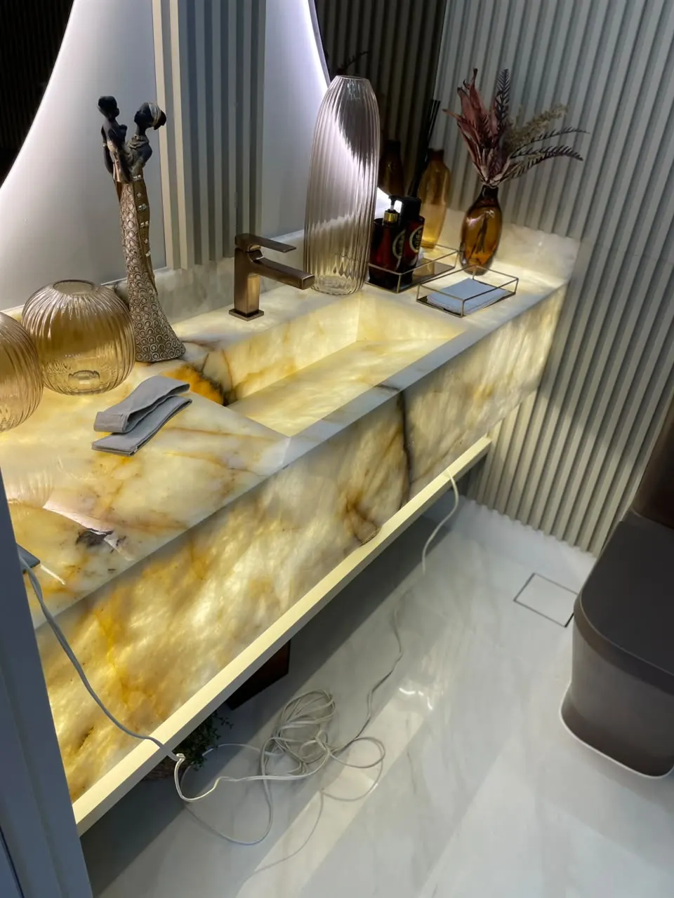 Bancada de pedra translúcida iluminada por dentro, com cuba esculpida e painel ripado ao fundo